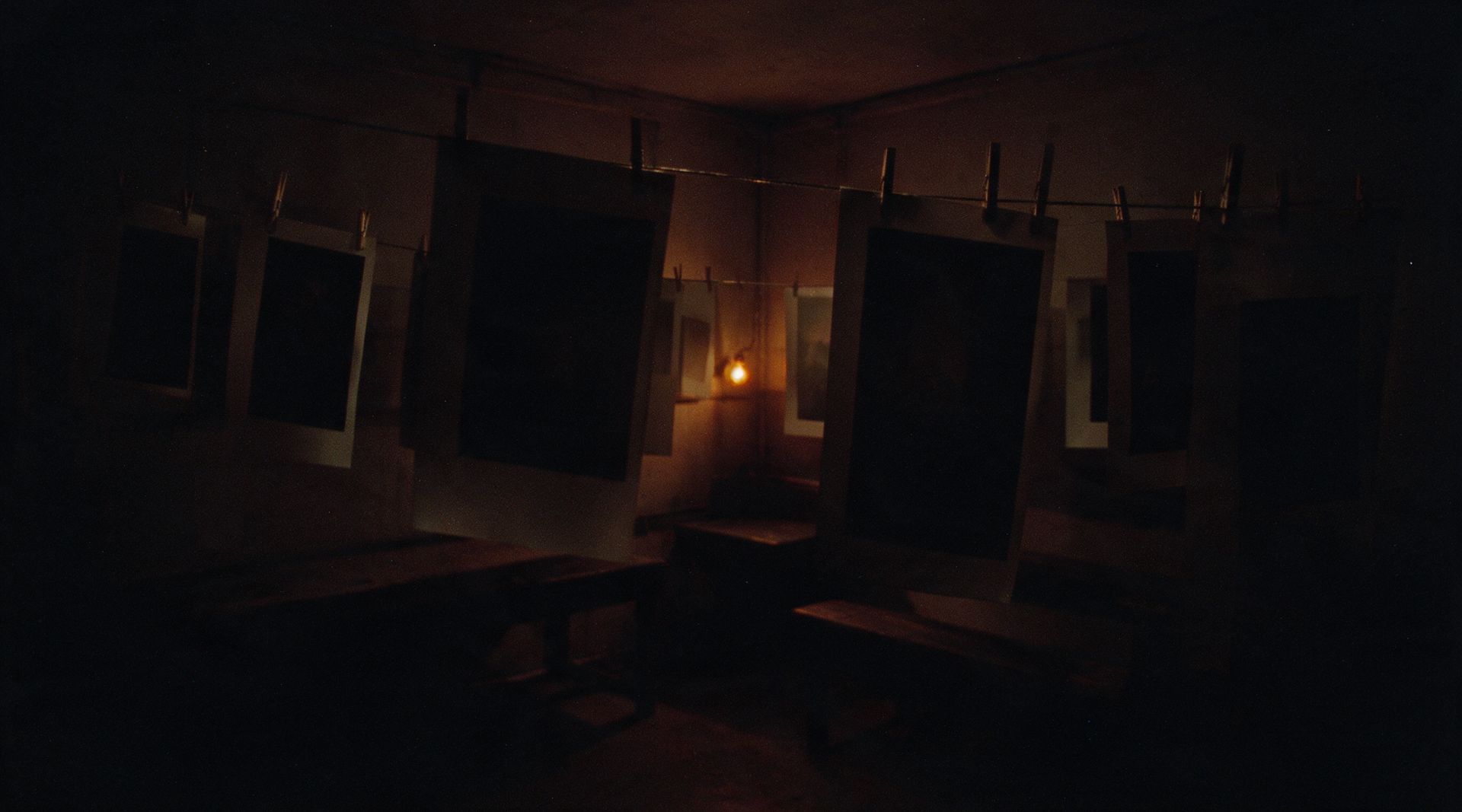 A photographic darkroom at night: blank prints hanging on a line under a single distant amber safelight — none of them developed yet.